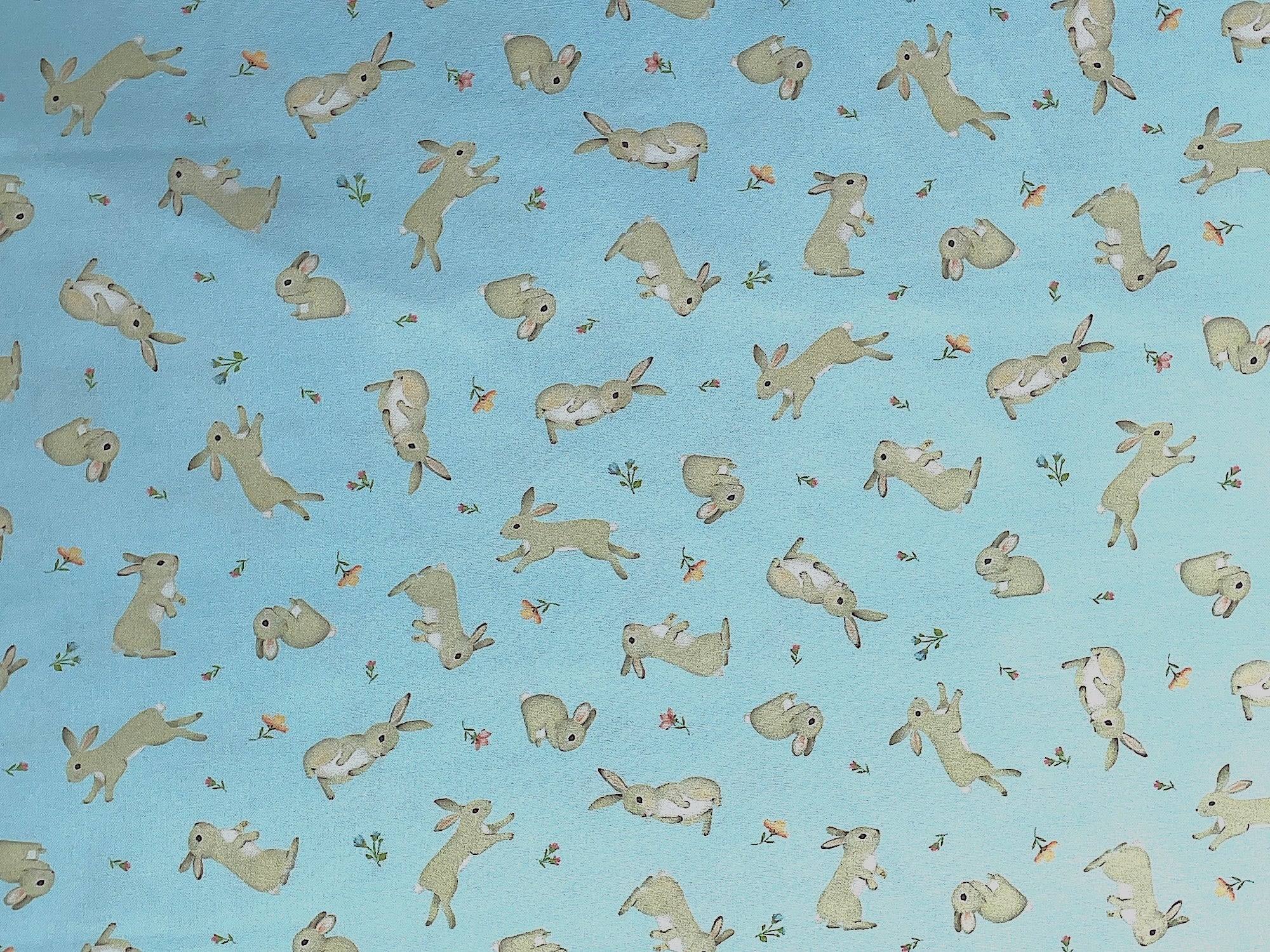 This light blue cotton fabric is covered with bunnies and flowers. the flowers are peach, blue and pink.