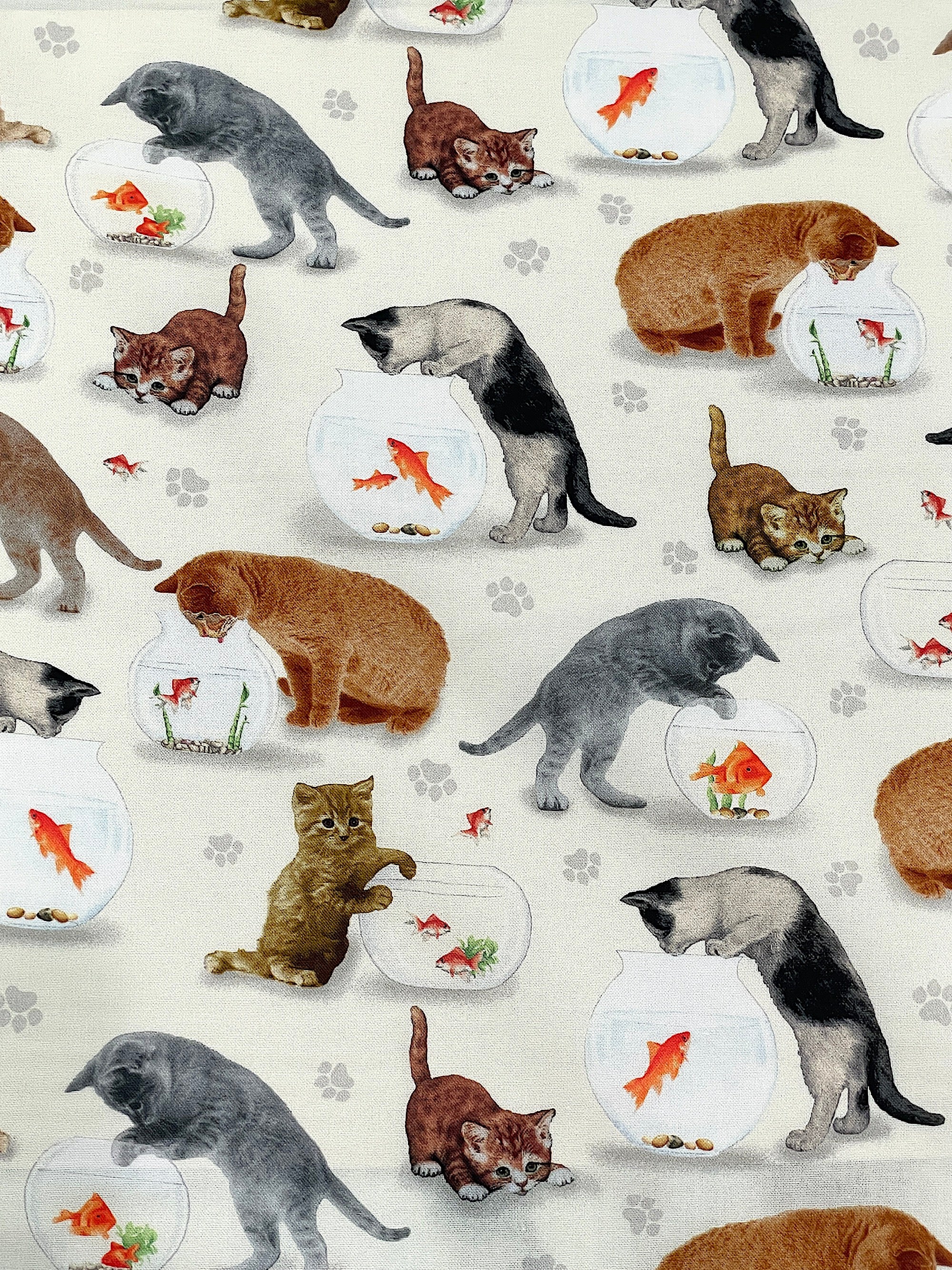 This cream colored cotton fabric is covered with cats and bowls of fish. The cats are a variety of colors such as grey, beige, brown and orange and they are either looking in at the fish or putting their paws in like they are trying to get a fish snack