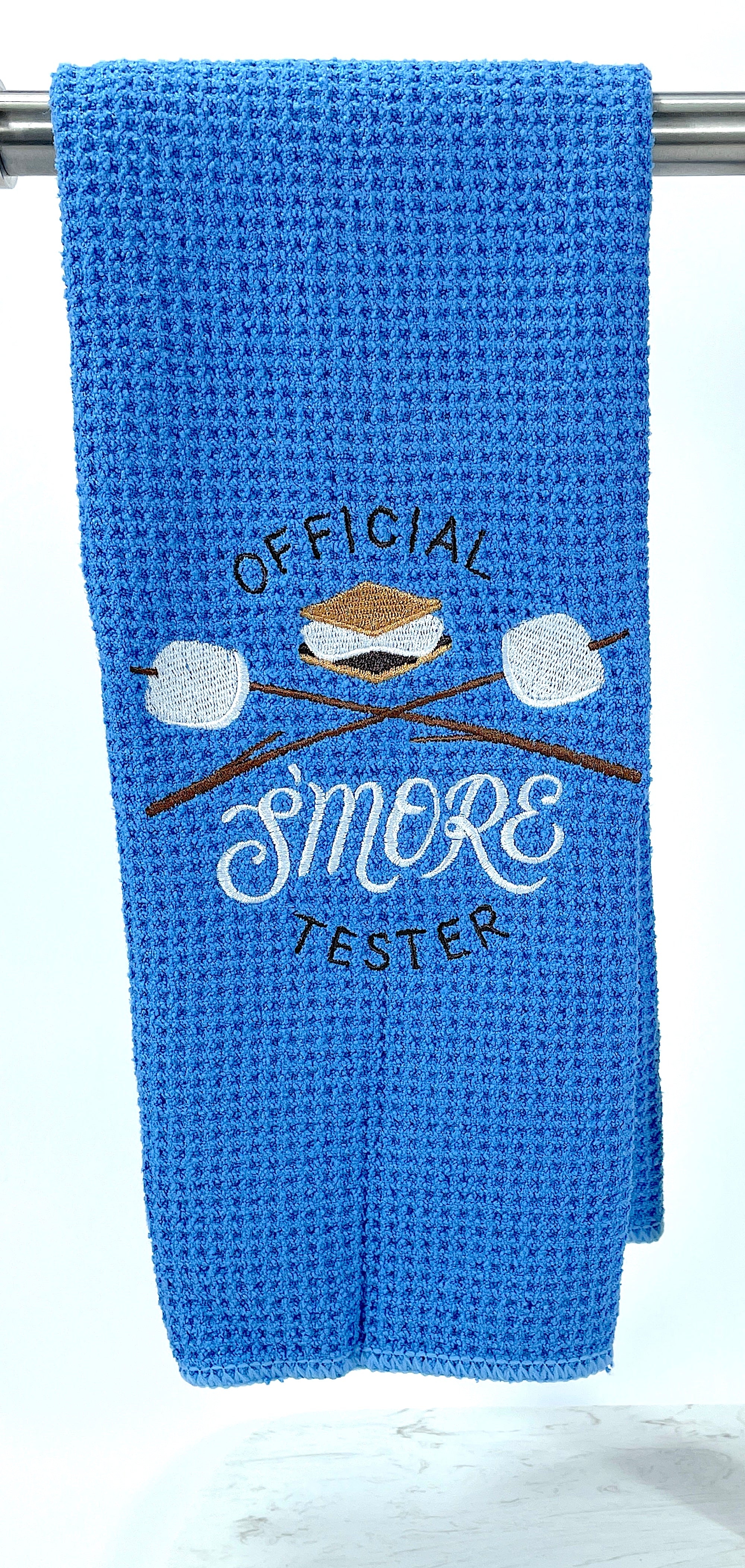 A blue microfiber waffle weave hand towel with 'OFFICIAL SMORE TESTER' embroidered on it, along with an image of a smore and marshmallows on sticks.