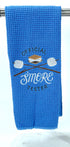 A blue microfiber waffle weave hand towel with 'OFFICIAL SMORE TESTER' embroidered on it, along with an image of a smore and marshmallows on sticks.