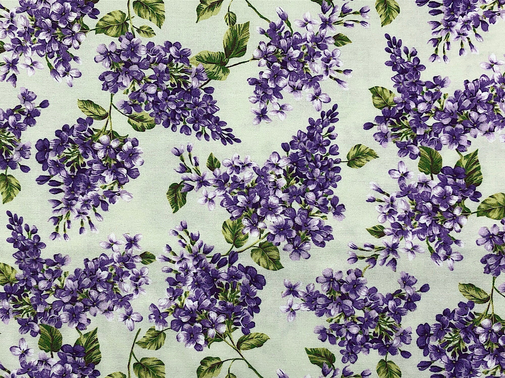 This white fabric is covered with Purple Lilacs and green leaves. The Lilacs are randomly placed throughout the pattern.