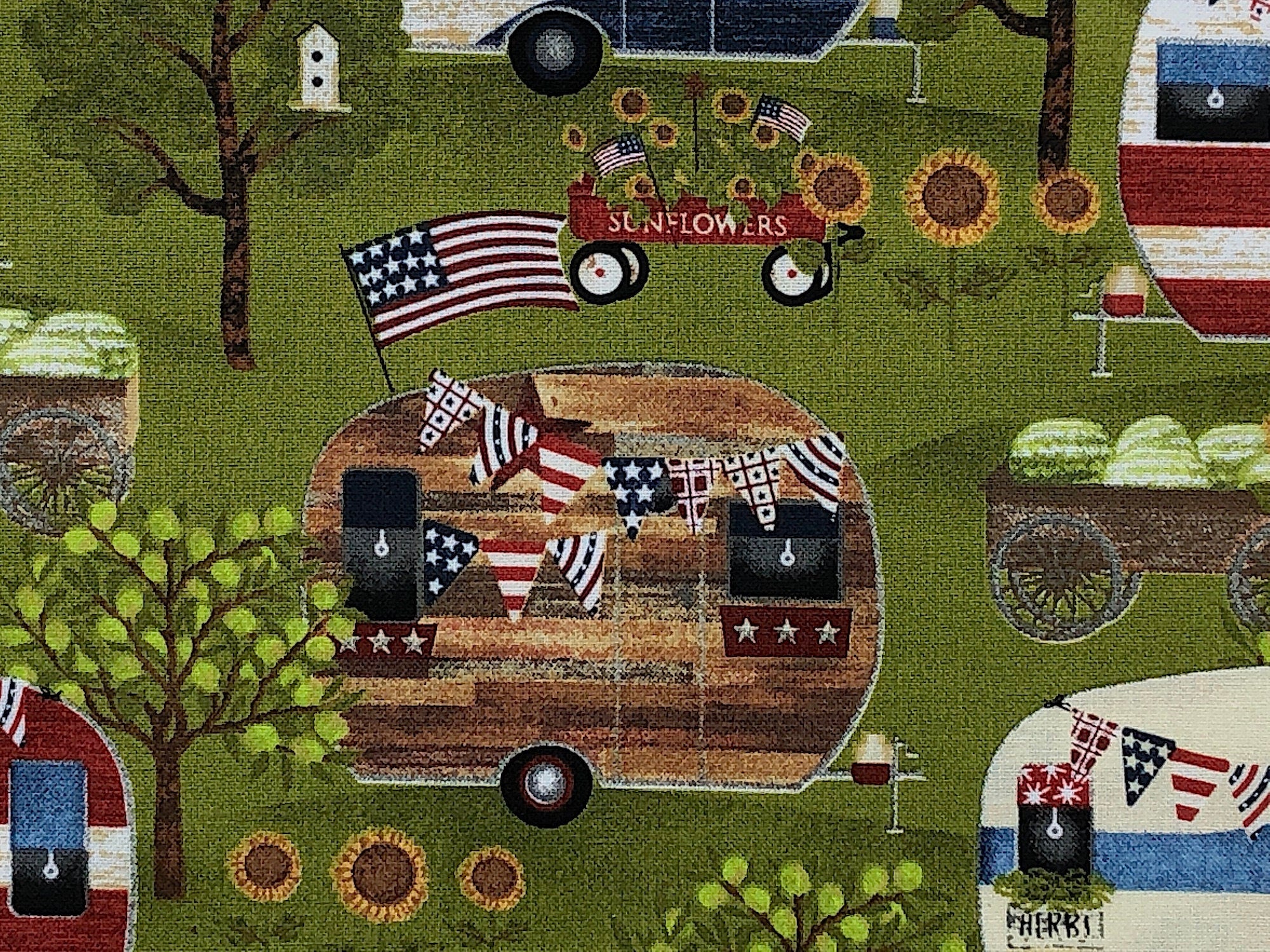 Close up of a brown travel trailer, trees, USA flag and wagon of flowers.