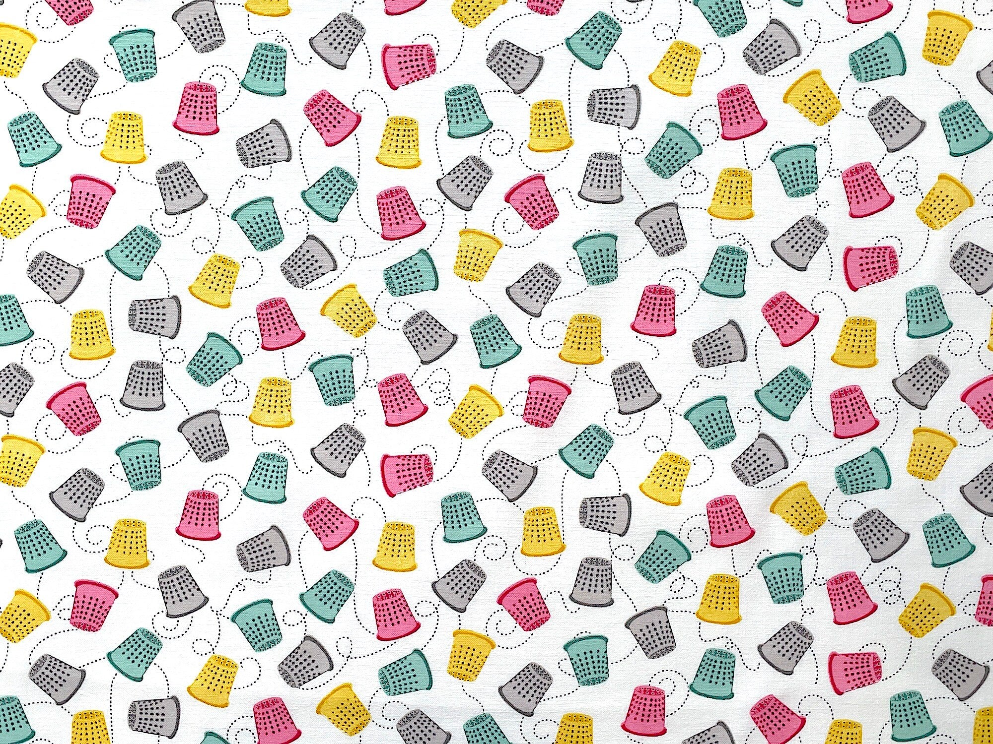 Green, yellow, pink and grey thimbles cover this white fabric. This fabric is part of the Words to Quilt by collection.