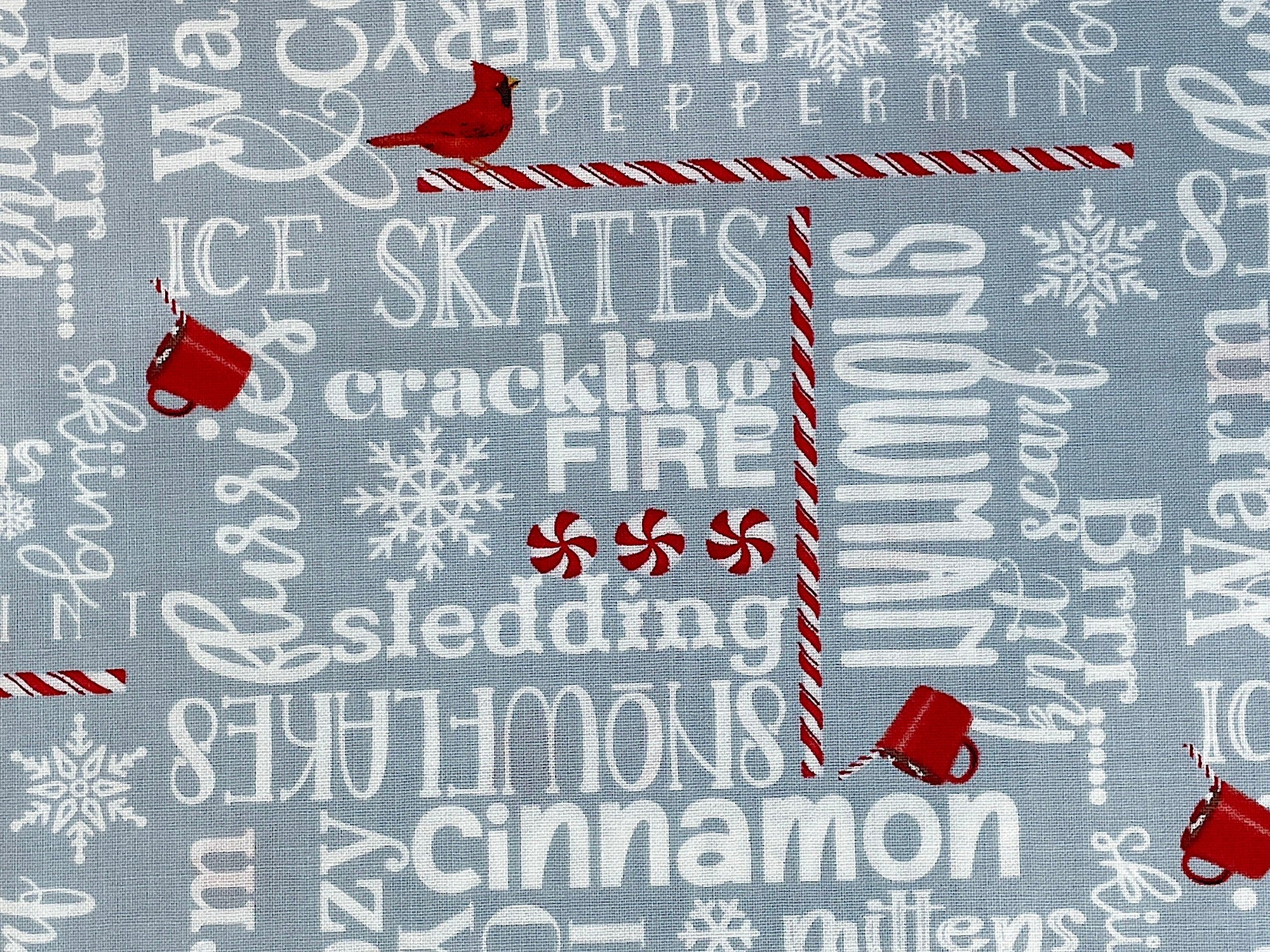 This gray cotton fabric is covered with winter words such as snowman, cinnamon, mittens, peppermint, crackling fire, ice skates and more. You will also find small red birds and red cups of cocoa throughout the fabric along with peppermint sticks.