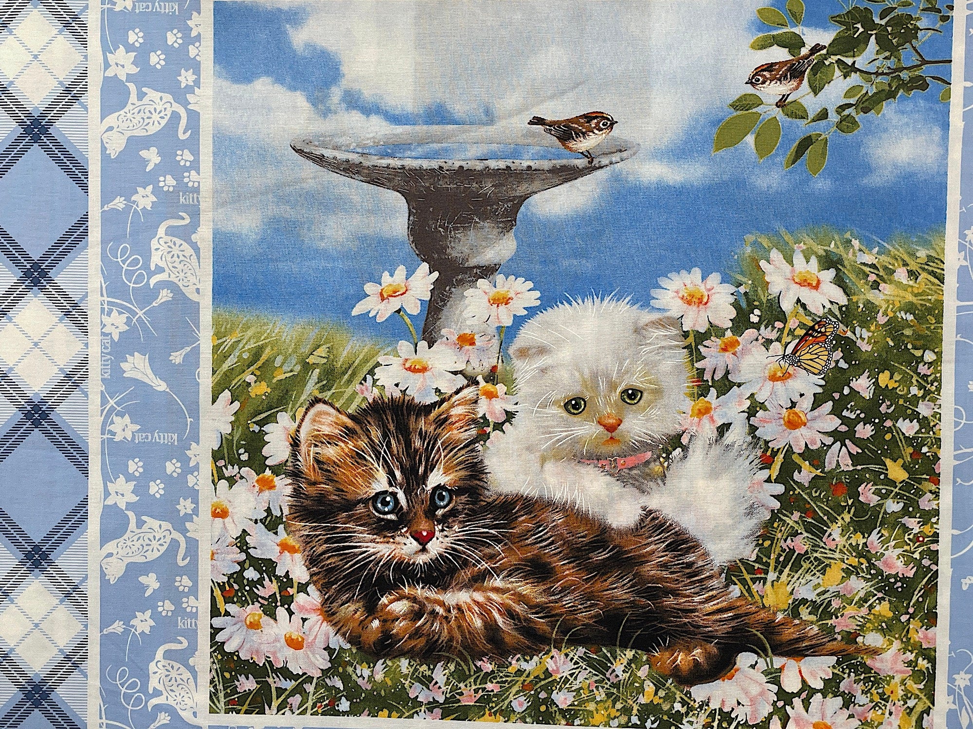Two cats laying in the grass with a birdbath behind them.