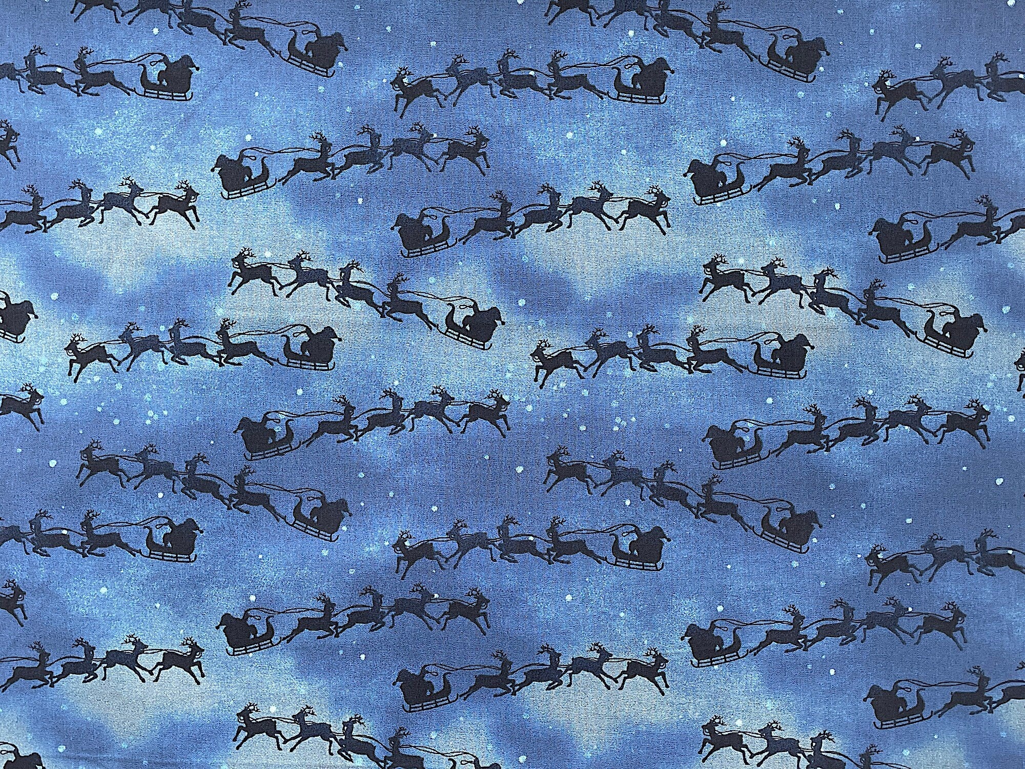 Santa and his reindeer are flying in the sky on this cotton fabric. Santa and his reindeer are black and the background has shades of blue and light tan.