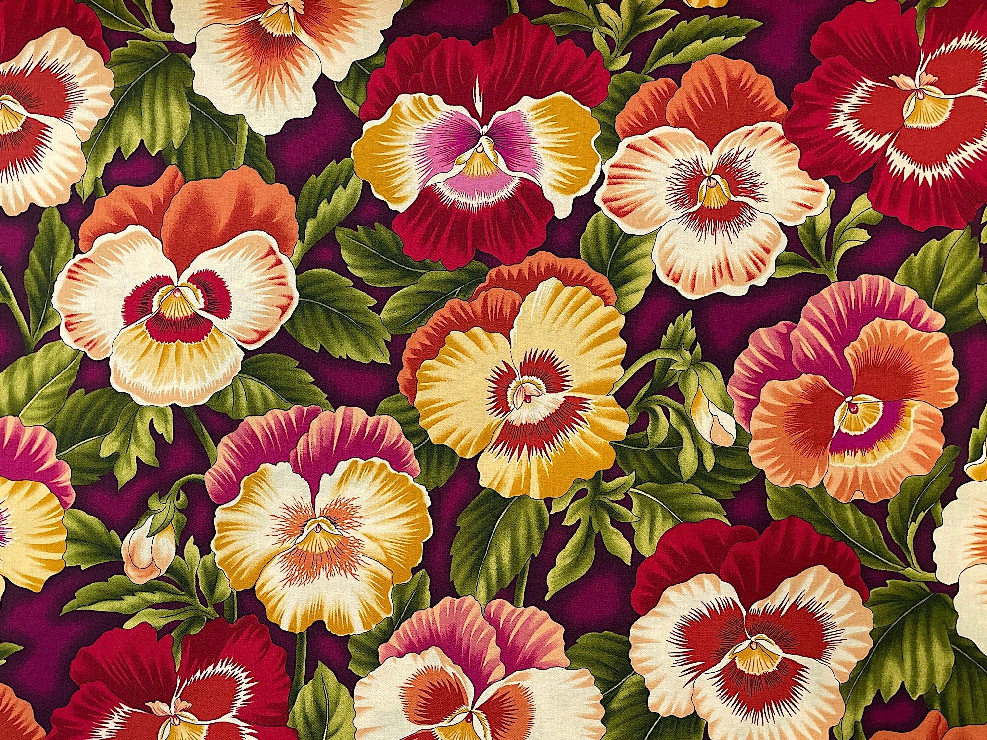 Cotton fabric covered with yellow, red, orange and white pansies and green leaves.