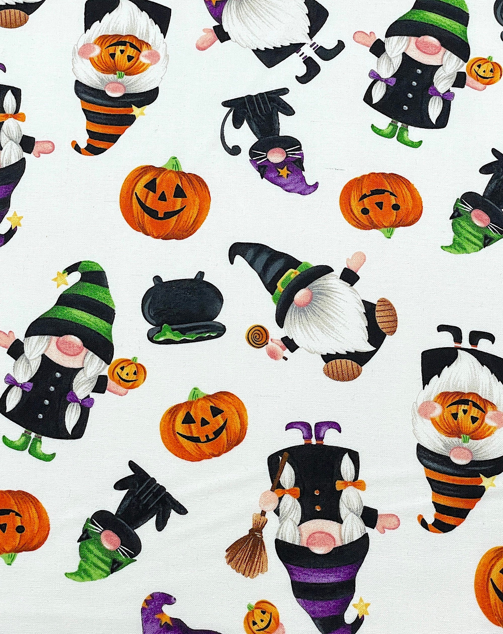 This white cotton fabric is covered with gnomes, black cats, kettles and pumpkins. The gnomes are holding pumpkins or brooms.